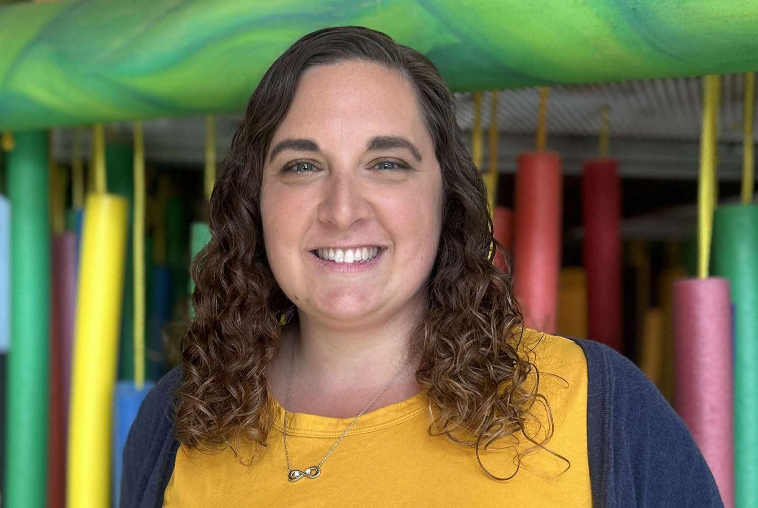 A woman with curly brown hair smiling, wearing a yellow shirt and a necklace with an infinity symbol pendant, standing in front of colorful playground equipment.