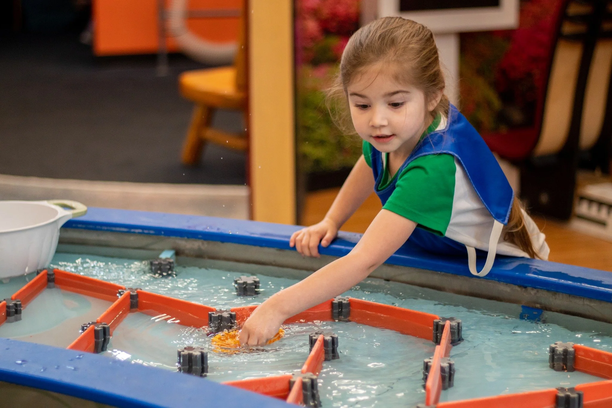 A young girl with red hair and wearing a blue apron over a green shirt plays at a water table in a classroom setting.