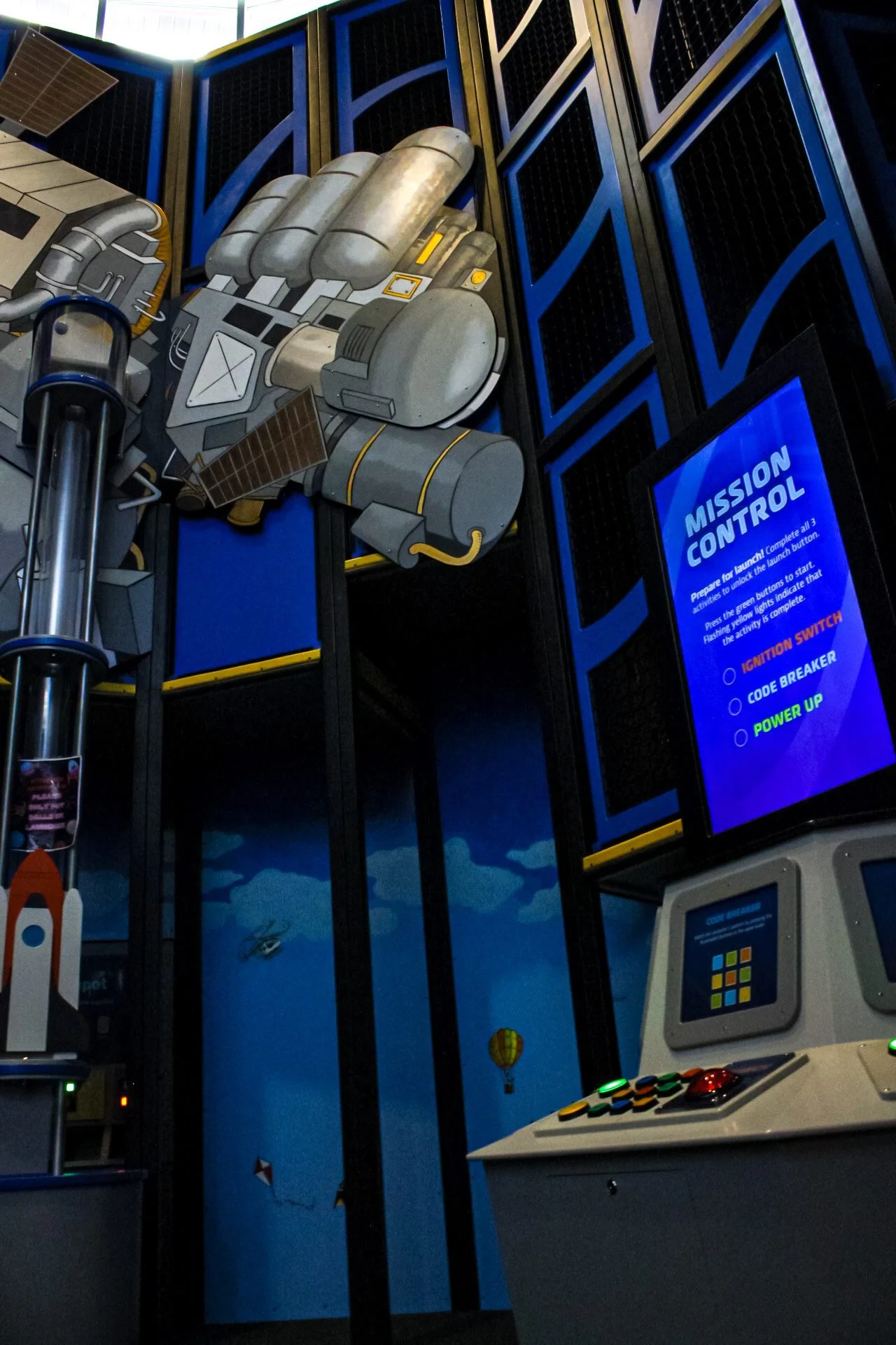 A large, cartoon-style depiction of a spacecraft's hand with buttons and electronic components is mounted on a wall. Next to it is a digital screen with instructions for a mission control game, and a control panel with colorful buttons and a joystick.