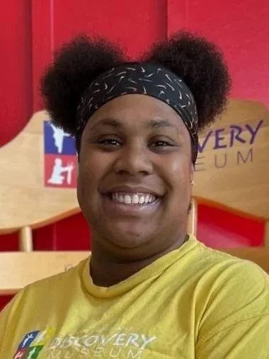 Smiling woman with dark skin, wearing a black bandana with white patterns and a yellow Discovery Museum t-shirt, in front of a red and yellow background.