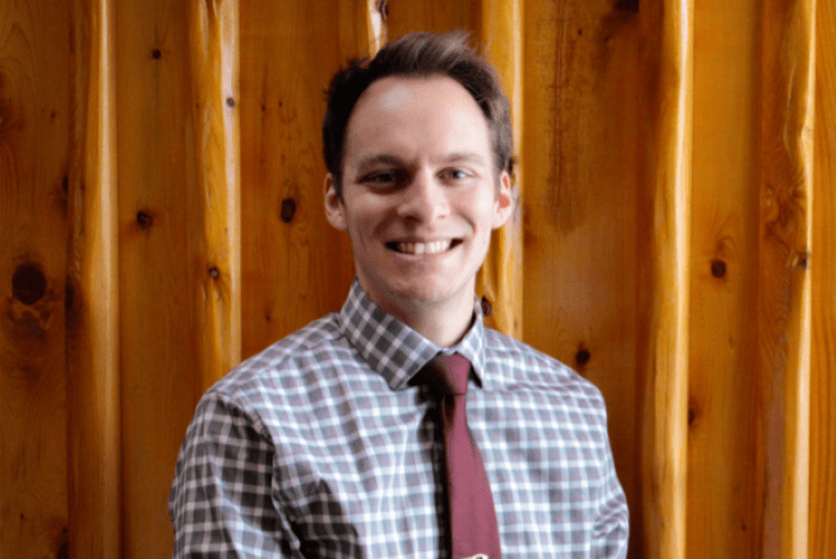 A smiling man with dark hair, wearing a checkered shirt and a maroon tie, standing in front of a wooden wall.
