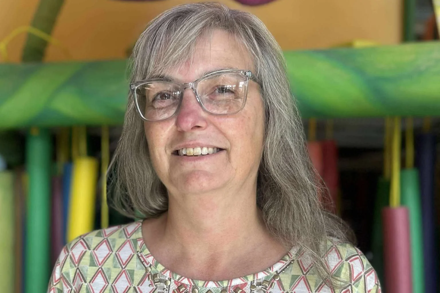A woman with gray hair wearing glasses and a patterned shirt, standing in front of colorful equipment or playground structures.