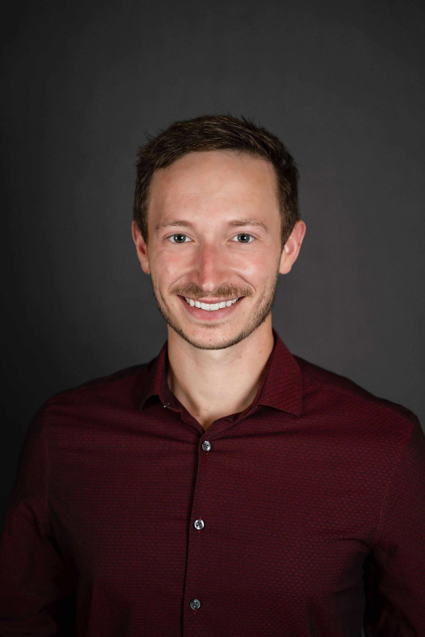 Portrait of a young man with short brown hair and a beard, smiling, wearing a maroon button-up shirt, against a dark background.