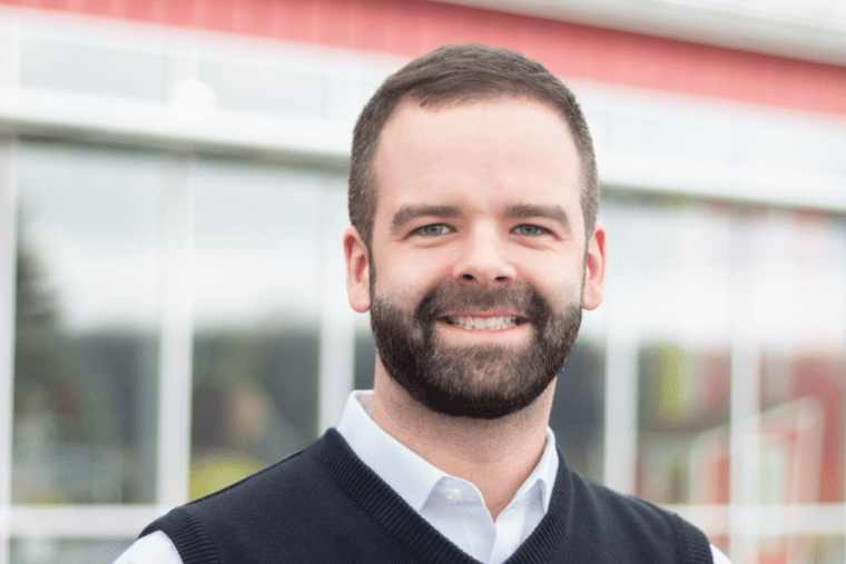 A man with a beard and short hair smiling outdoors in front of a modern building with glass windows.