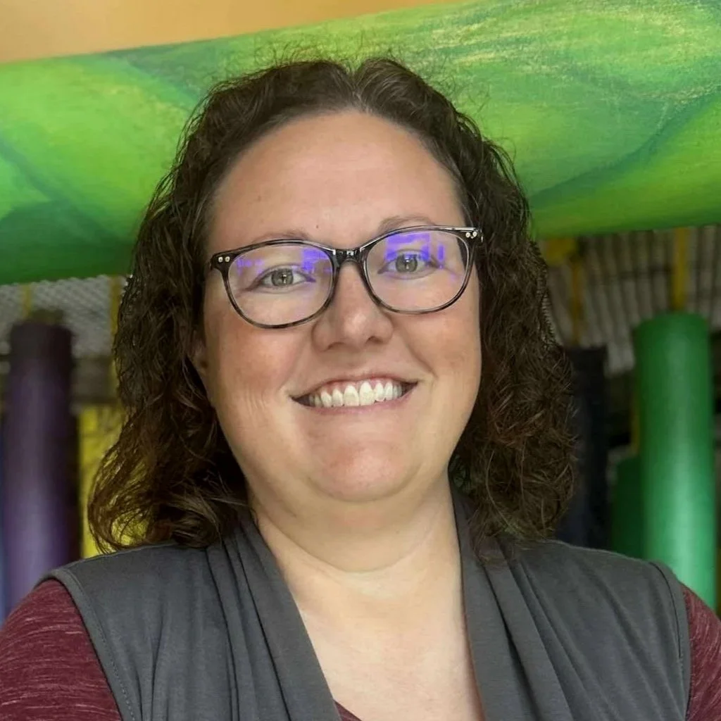 Smiling woman with glasses in indoor play area with colorful foam tubes and structures in the background.