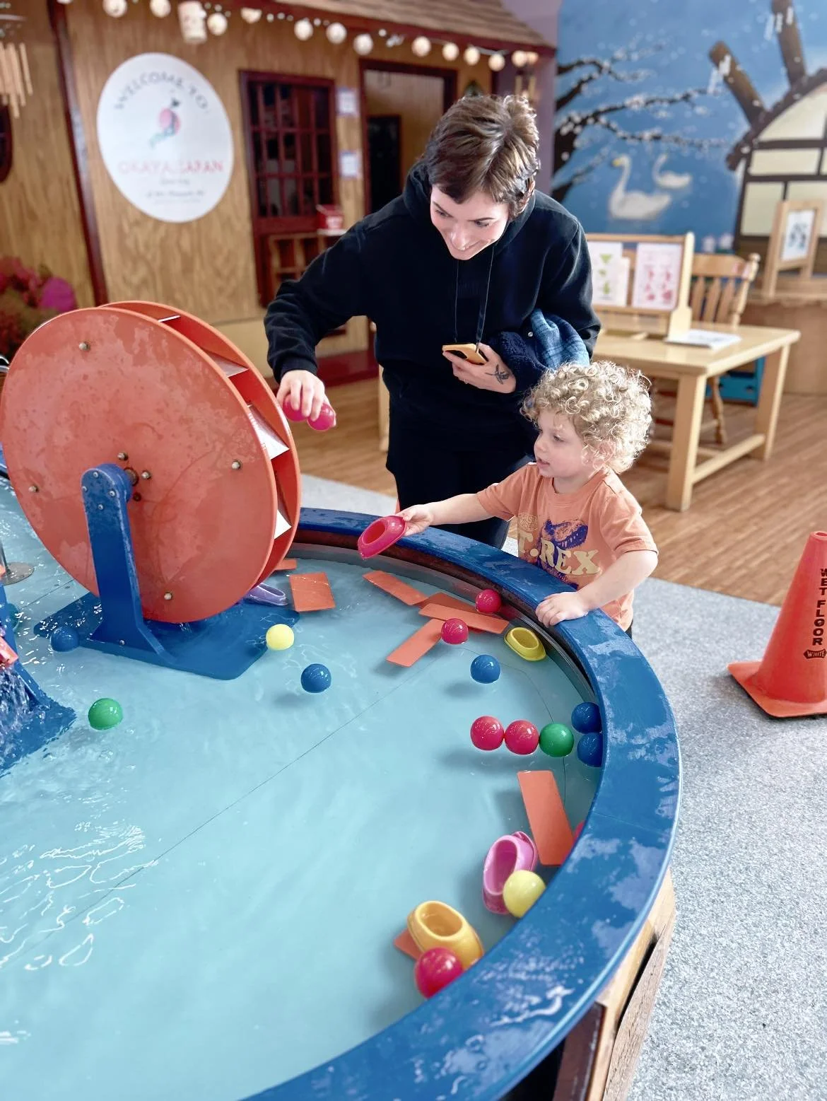 A woman and a young child playing an indoor pinball game with colorful balls and paddles, in a room with wood-paneled walls and decorative murals.