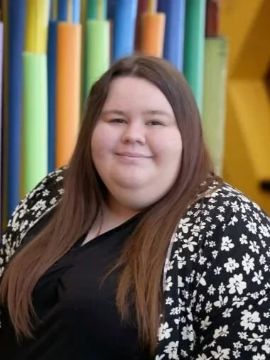 A young woman with long brown hair smiling in front of colorful foam pool noodles.