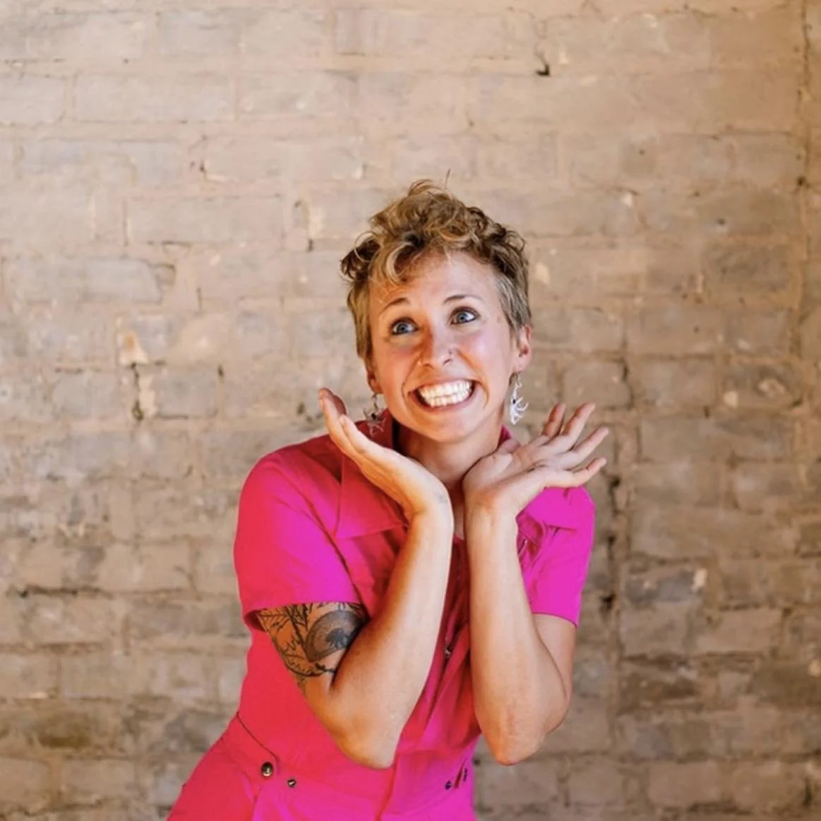 A woman with short hair and tattoos on her arm, wearing a bright pink shirt, smiling widely with hands near her face, standing against a brick wall background.