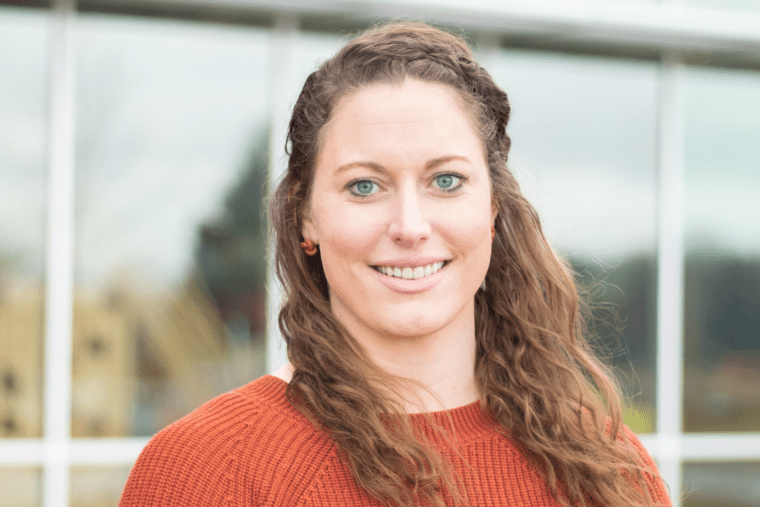 A woman with long, curly brown hair and blue eyes, wearing a red sweater, standing outdoors in front of a building with glass windows.