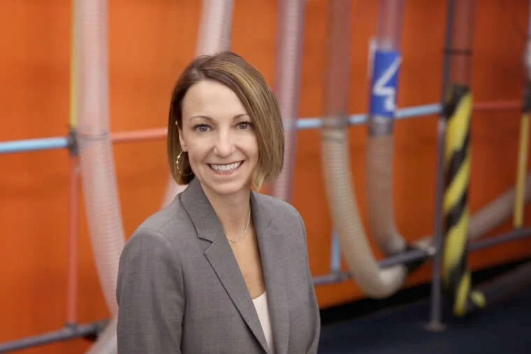 Smiling woman in a gray blazer standing in front of an industrial background with pipes and orange walls.