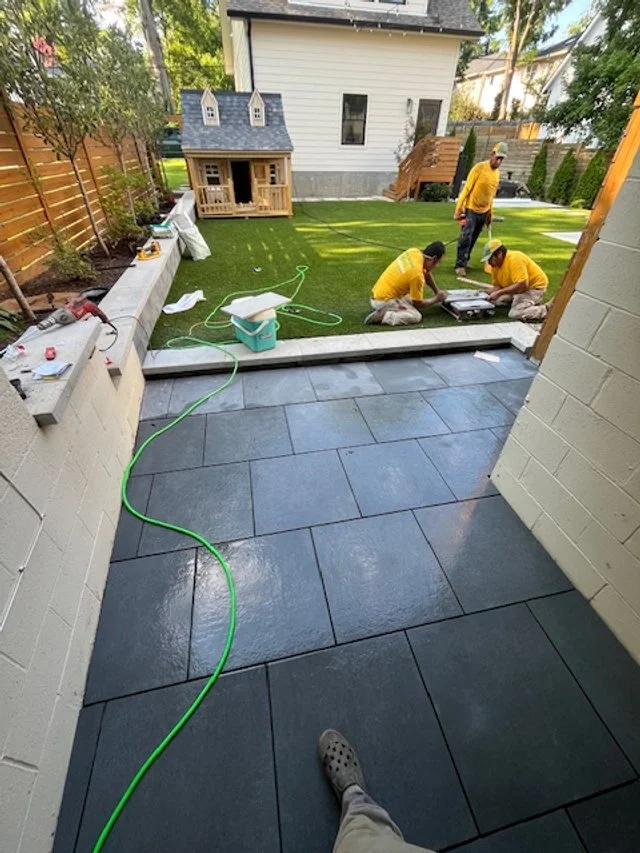 Workers installing large dark gray tiles on a patio next to a house, with tools and supplies nearby, in a backyard with a small house and trees.