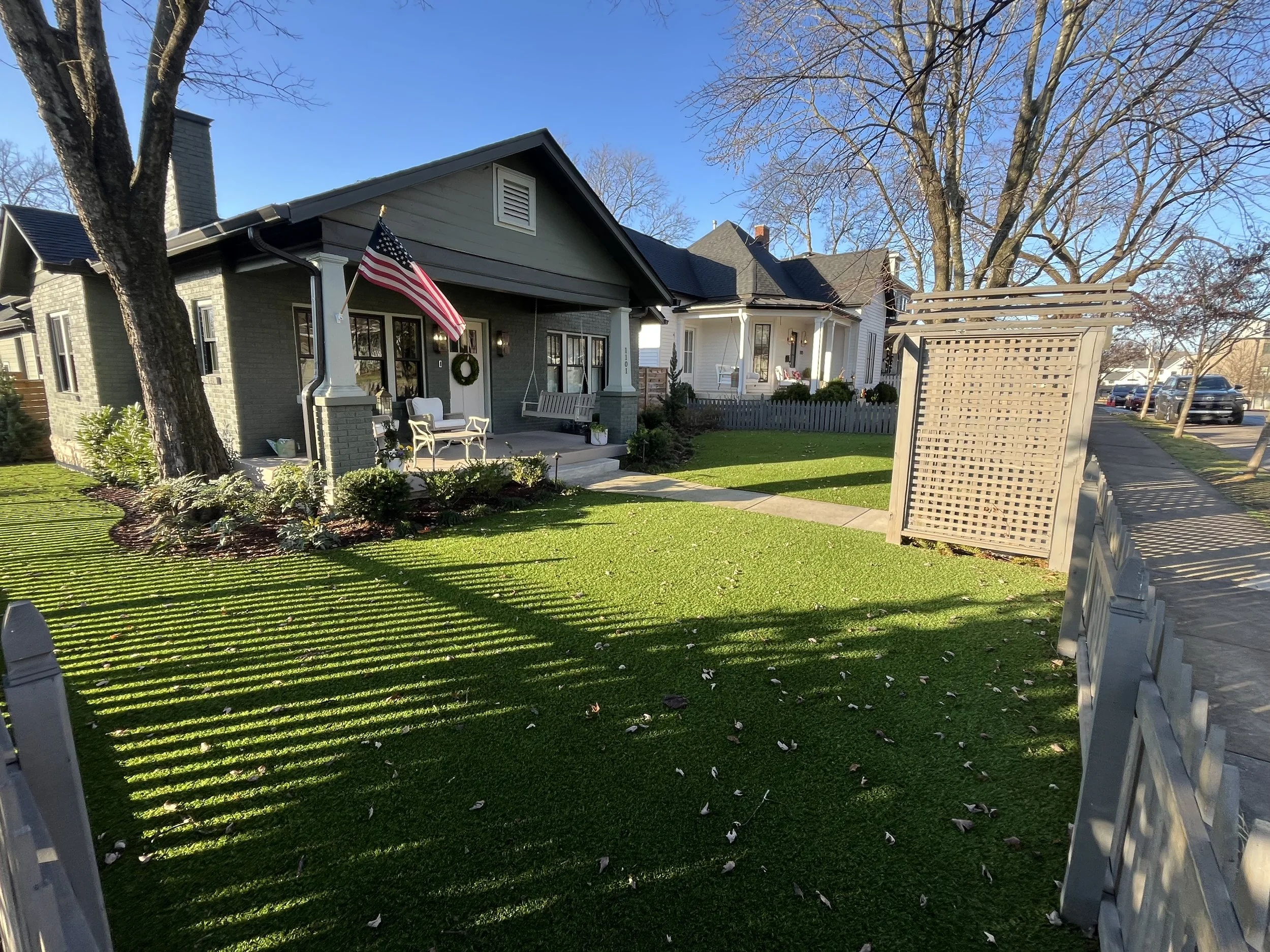 A house with a front porch decorated with an American flag and a wreath on the door, surrounded by a green lawn and trees, with a sidewalk and parked cars in the background.
