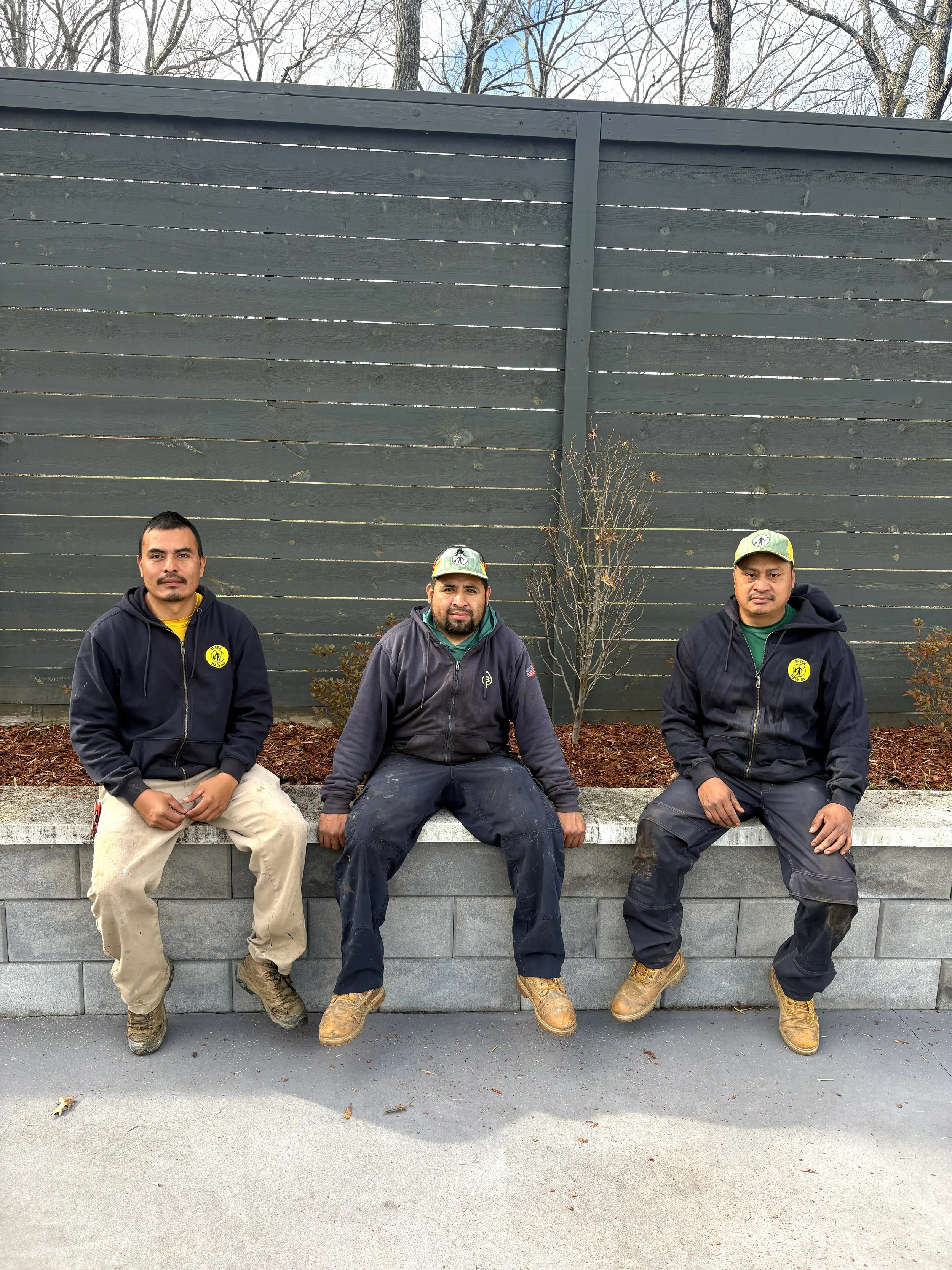 Three Green Machine employees sitting on a concrete ledge in front of a dark wooden fence with a small tree and brown shrubs behind them, wearing work clothing and boots.