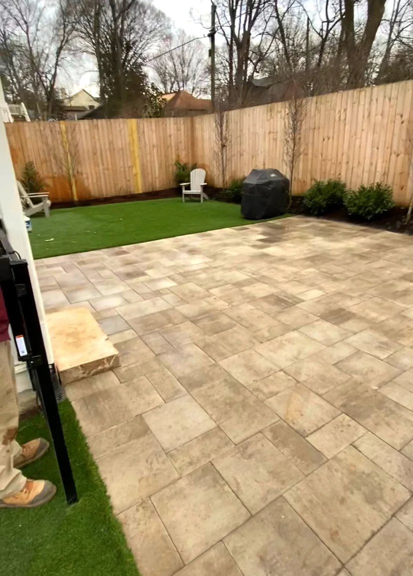 A backyard with a paved patio area, a small grassy section with white chairs, a black grill covered with a tarp, and a wooden fence surrounding the yard. There are a few small trees and shrubs along the fence line.