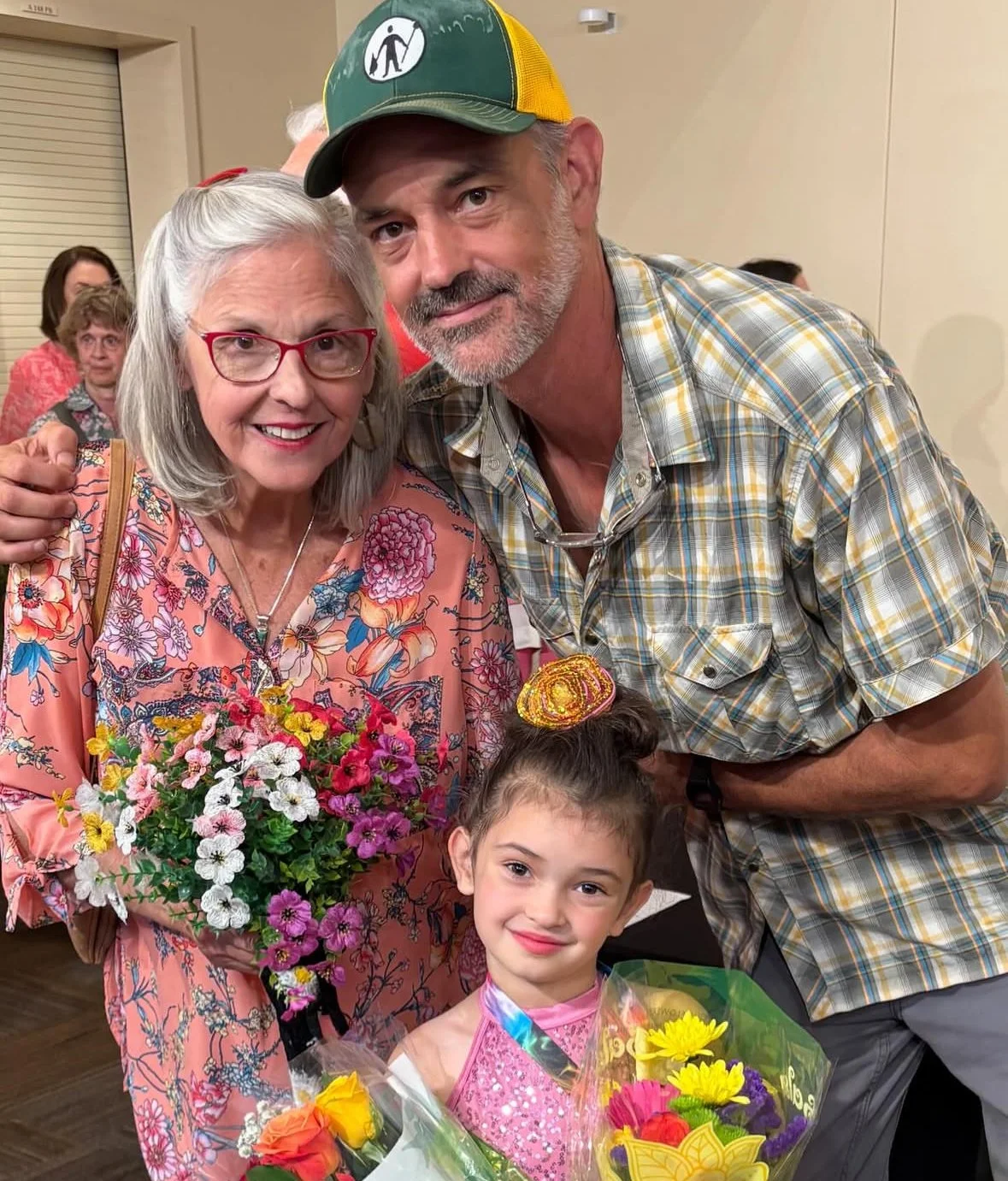 A group of four people, including an elderly woman with gray hair, glasses, and a bright floral dress holding a bouquet of flowers, a young girl with a pink dress and a colorful hair accessory holding a bouquet of flowers, and two men, one with a beard wearing a plaid shirt and the other wearing a cap, posing together at an indoor event.