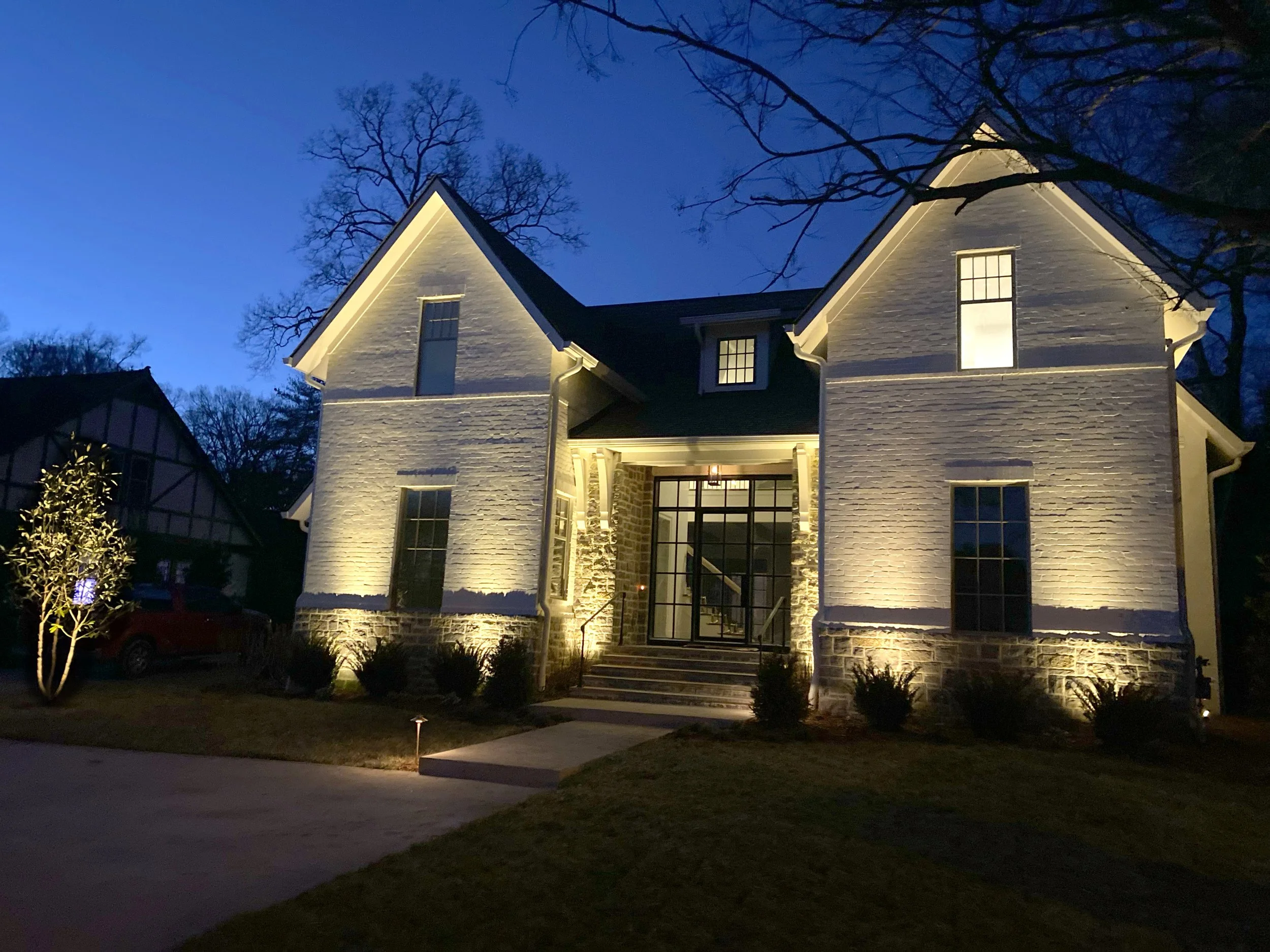 A two-story house at dusk, illuminated by exterior lighting, with a front porch, stairs, six windows, and a landscaped yard with small bushes and a tree with lights.