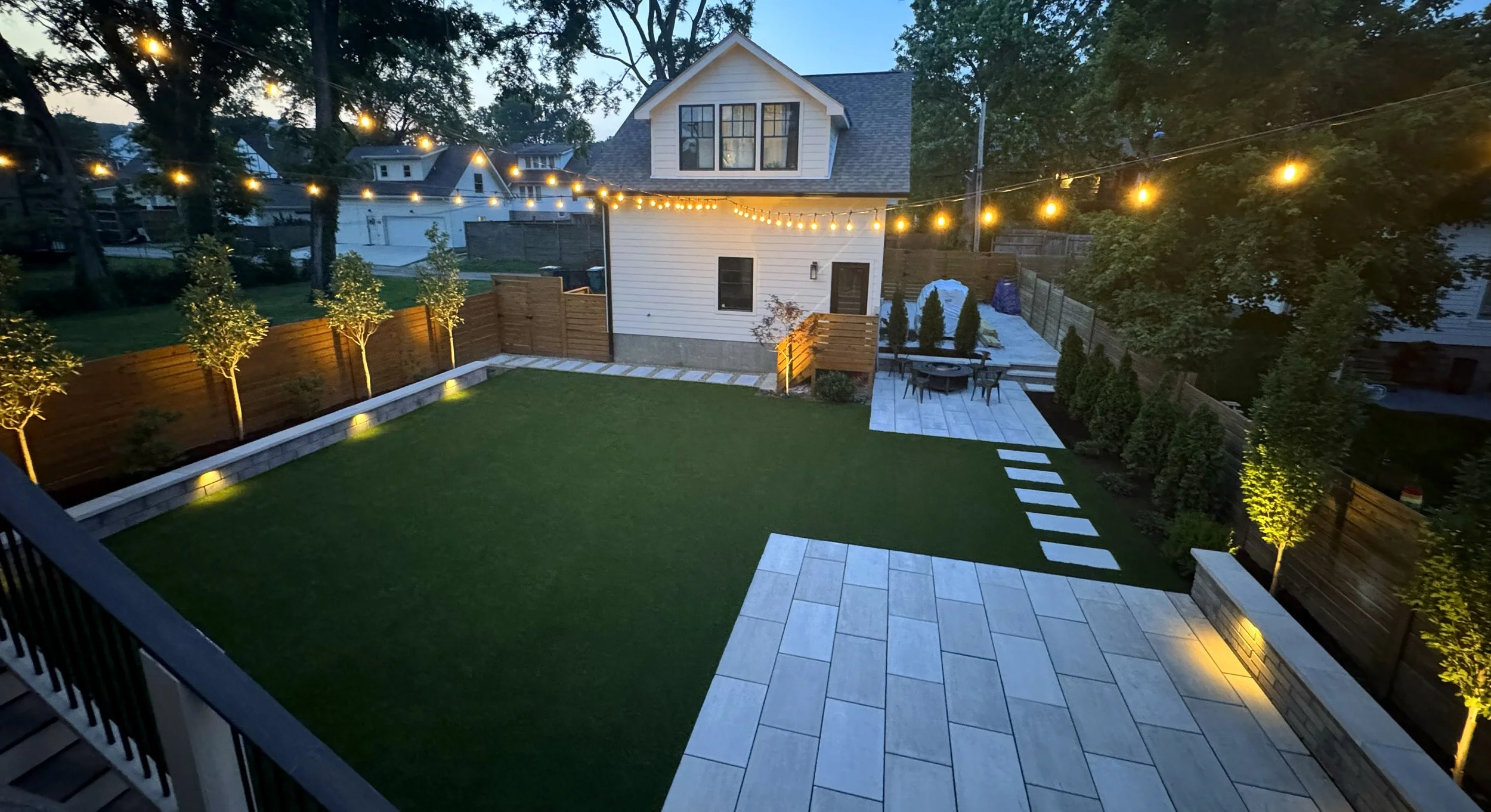 A backyard with a well-maintained lawn, trees with spotlights, a wooden fence, a sitting area with a table and chairs, string lights overhead, a paved patio area, and a white house in the background during twilight.