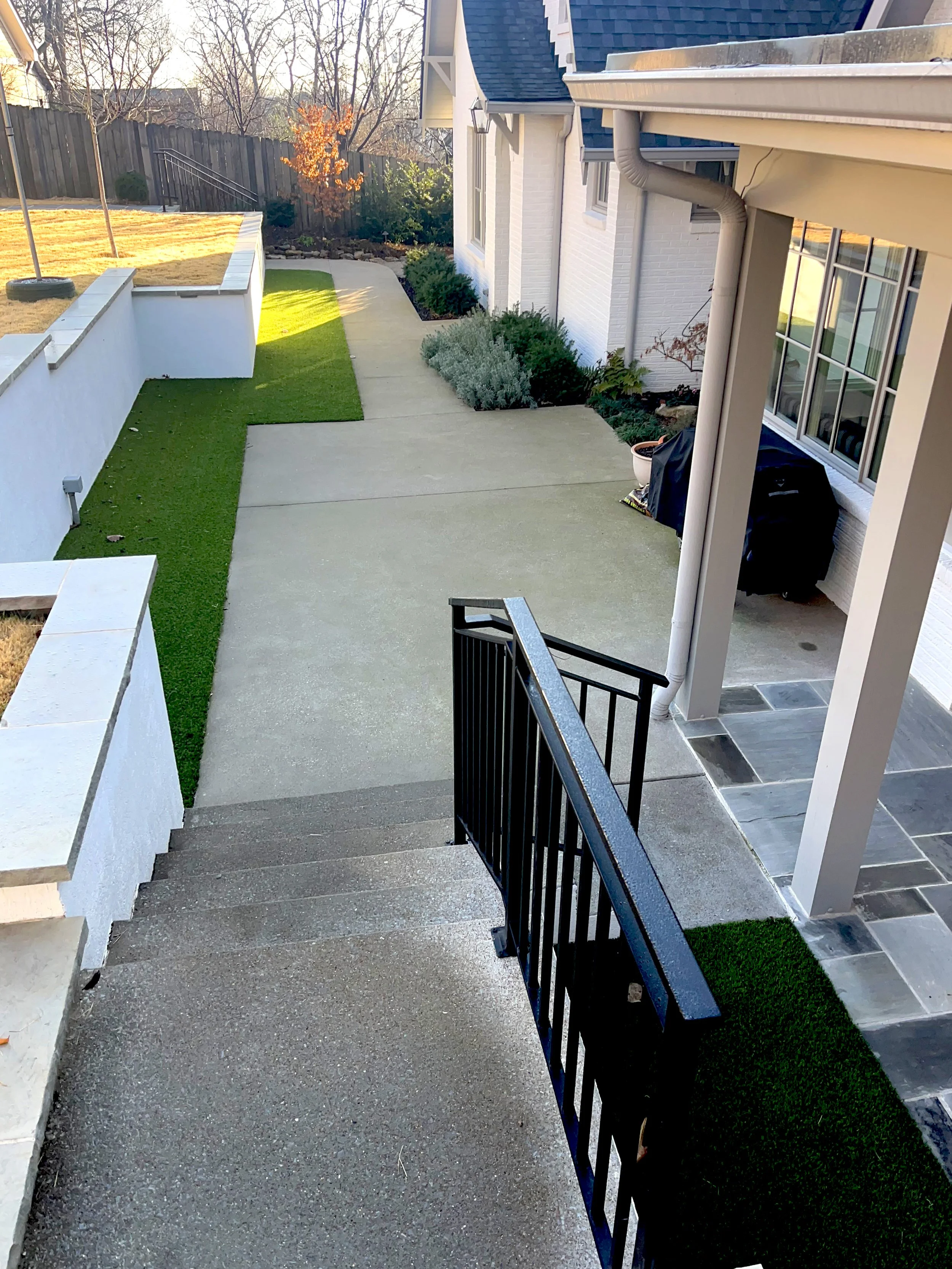 View of a backyard patio area with concrete stairs leading down to a concrete walkway, a small lawn, and a landscaped garden with bushes and a tree with orange leaves, fenced yard, and the side of a house with a sliding glass door, window, and exterior wall.