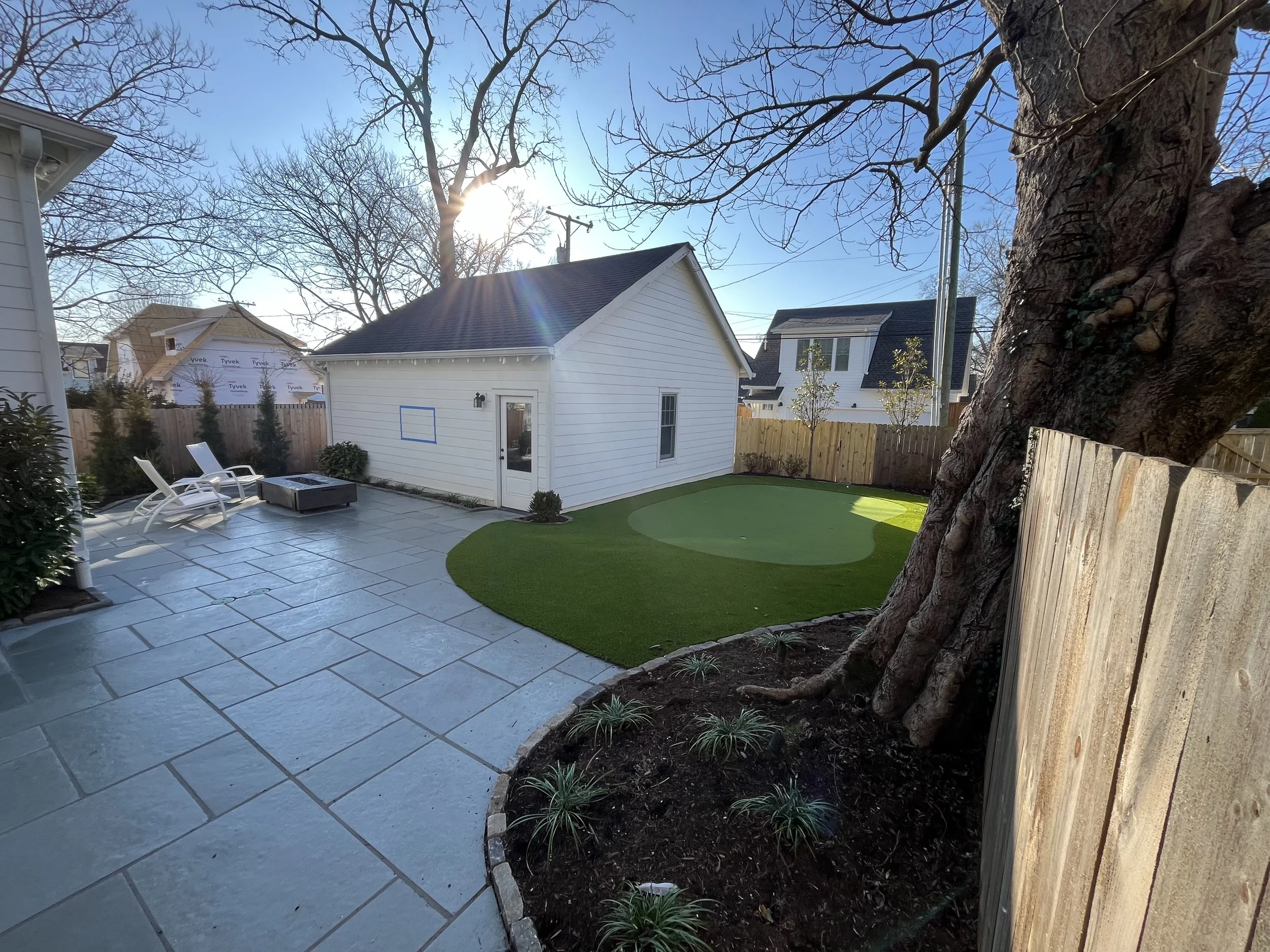 A backyard with a tiled patio, two white lounge chairs, a fire pit, a small white building, a patch of artificial turf, a large tree, a wooden fence, and neighboring houses in the background.