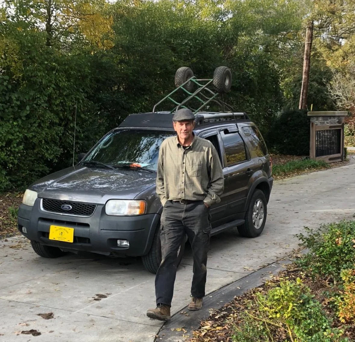 A man in casual clothing standing in front of a black SUV with a roof rack holding a green wheelbarrow. The scene takes place on a driveway with greenery and trees in the background.