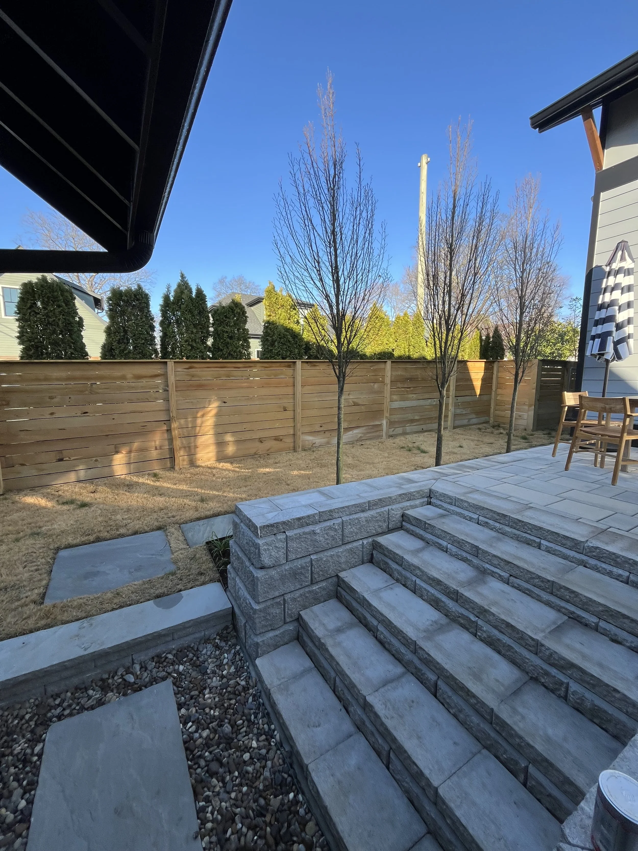 Backyard patio with stone steps leading down to a grassy yard, enclosed by a wooden fence, with leafless trees and neighboring houses in the background, under a clear blue sky.
