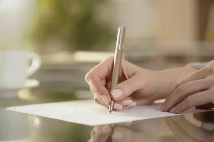 Close-up of a person's hand writing on a white sheet of paper with a silver pen, with a blurred background including a white coffee cup.