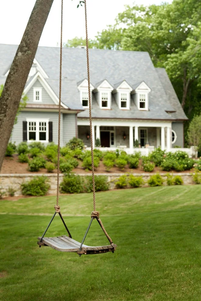 A wooden swing hanging from a tree branch in a backyard with a large house and well-maintained lawn and shrubs in the background.