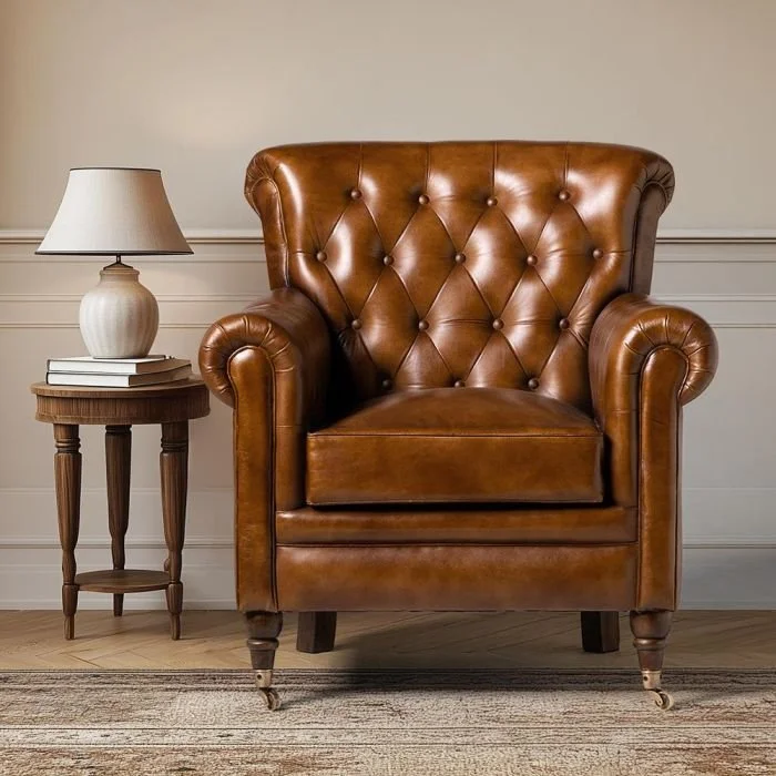 A leather armchair with a high, tufted back and rolled arms, placed next to a small wooden side table with a lamp and a few books in a living room setting.