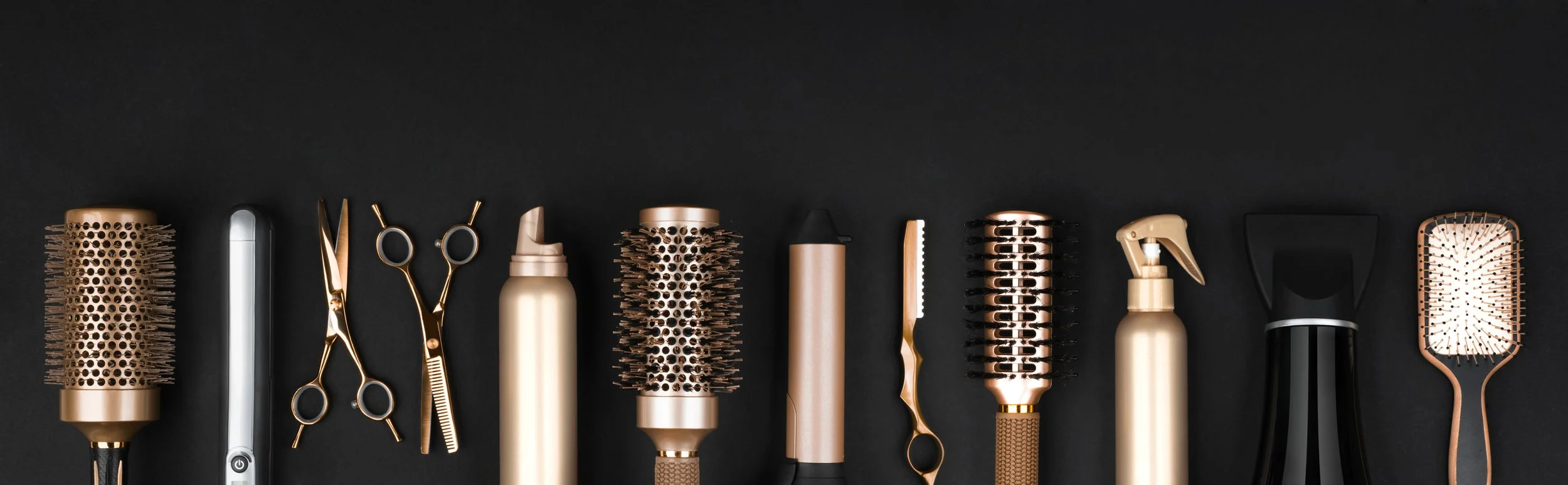 Collection of hair styling tools with gold accents arranged on a black background, including brushes, scissors, spray bottle, and clippers.