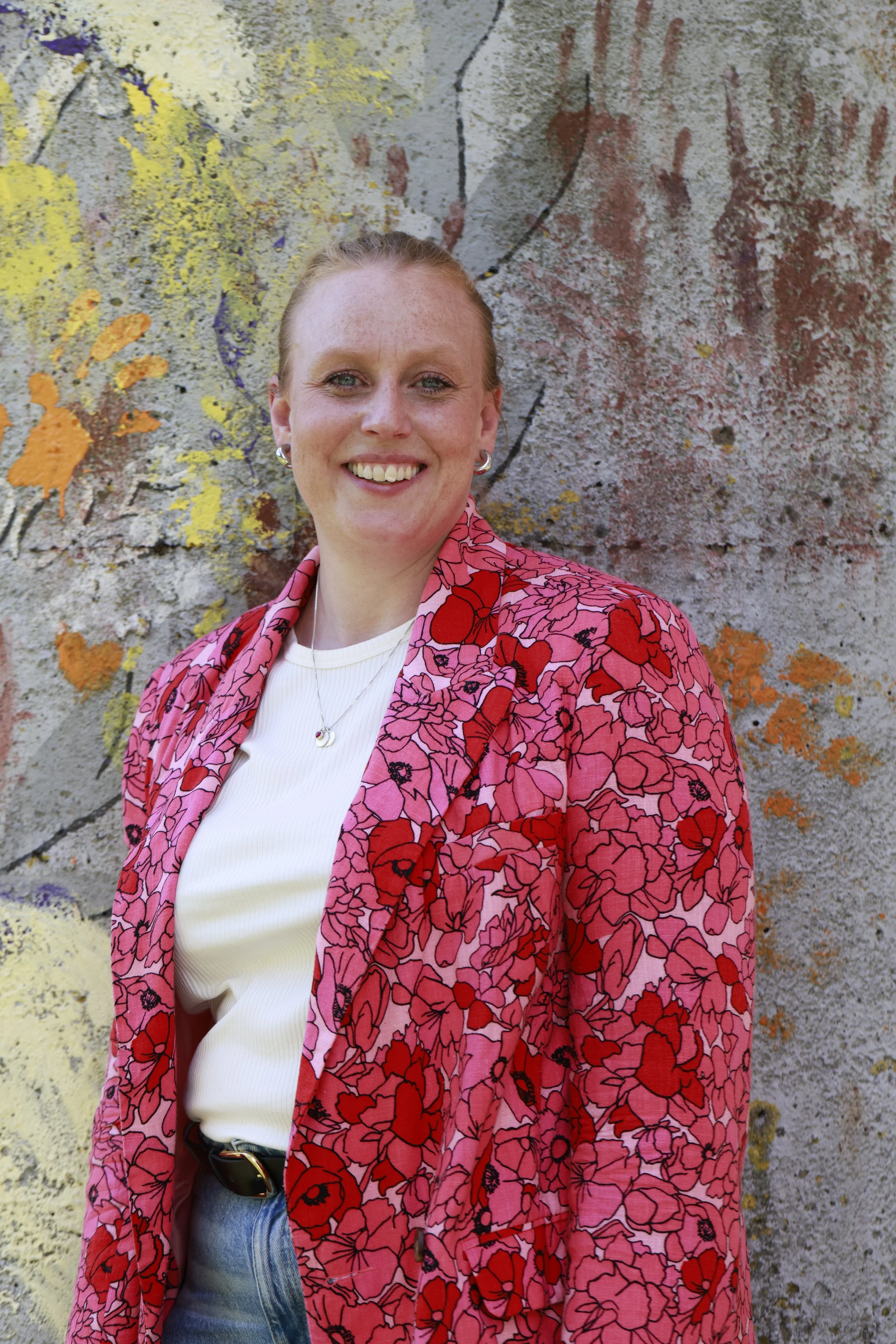 Mikaela with red hair smiling, standing outdoors in front of a colorful, graffiti-covered wall. She is wearing a pink and red floral jacket over a white top, with a necklace and earrings.