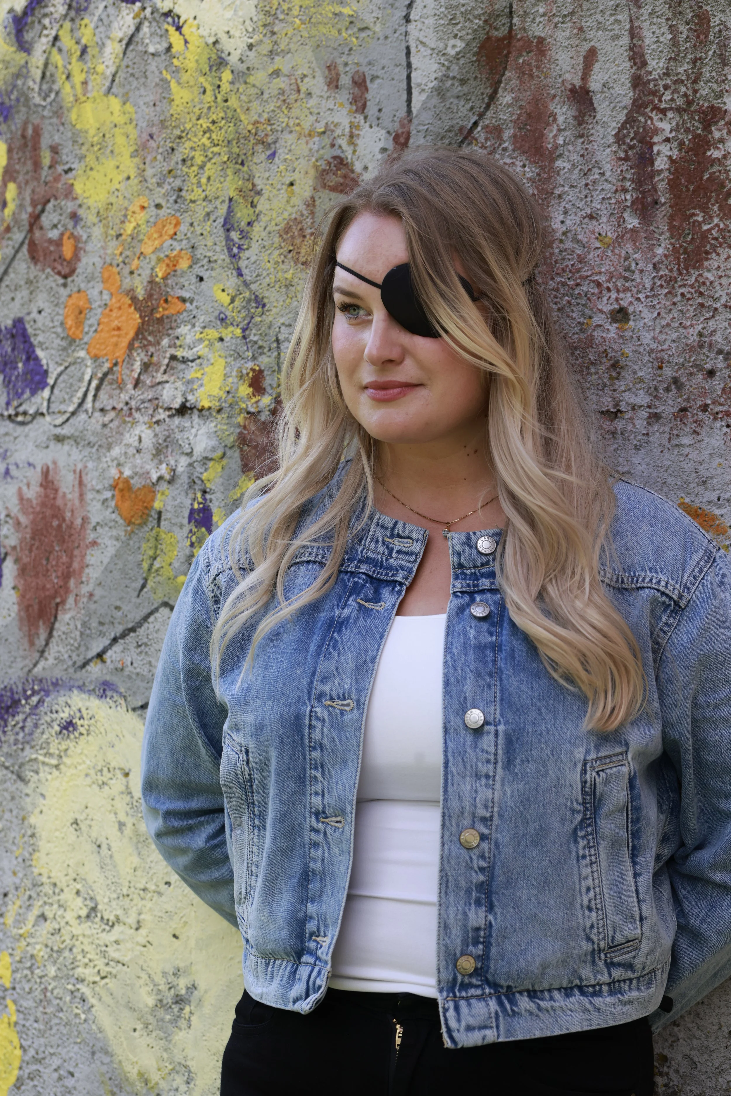 A woman with a black eye patch over her right eye, wearing a denim jacket and white top, standing in front of a graffiti-covered wall.