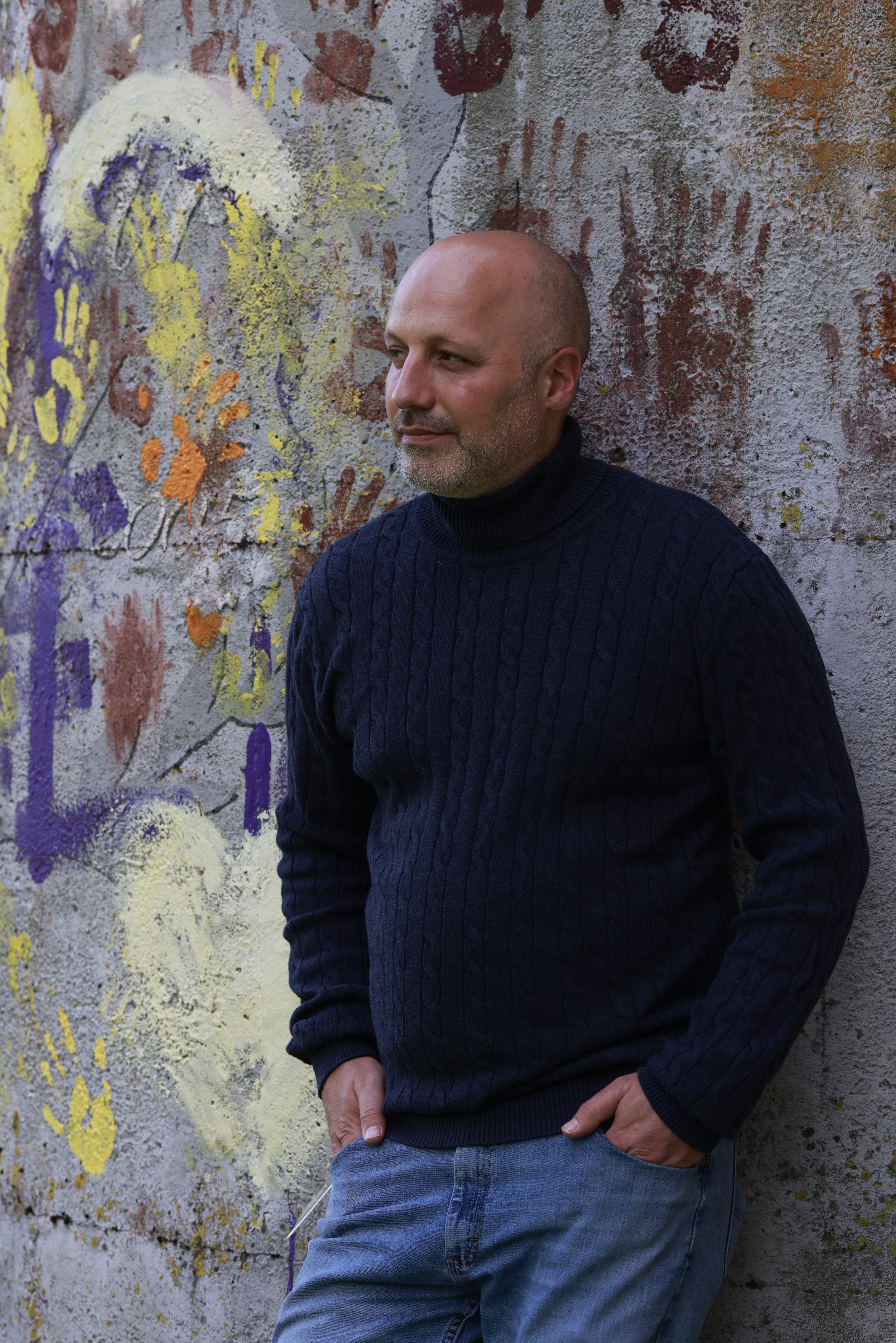A man with a shaved head and beard leaning against a wall with colorful handprints and spray paint art.