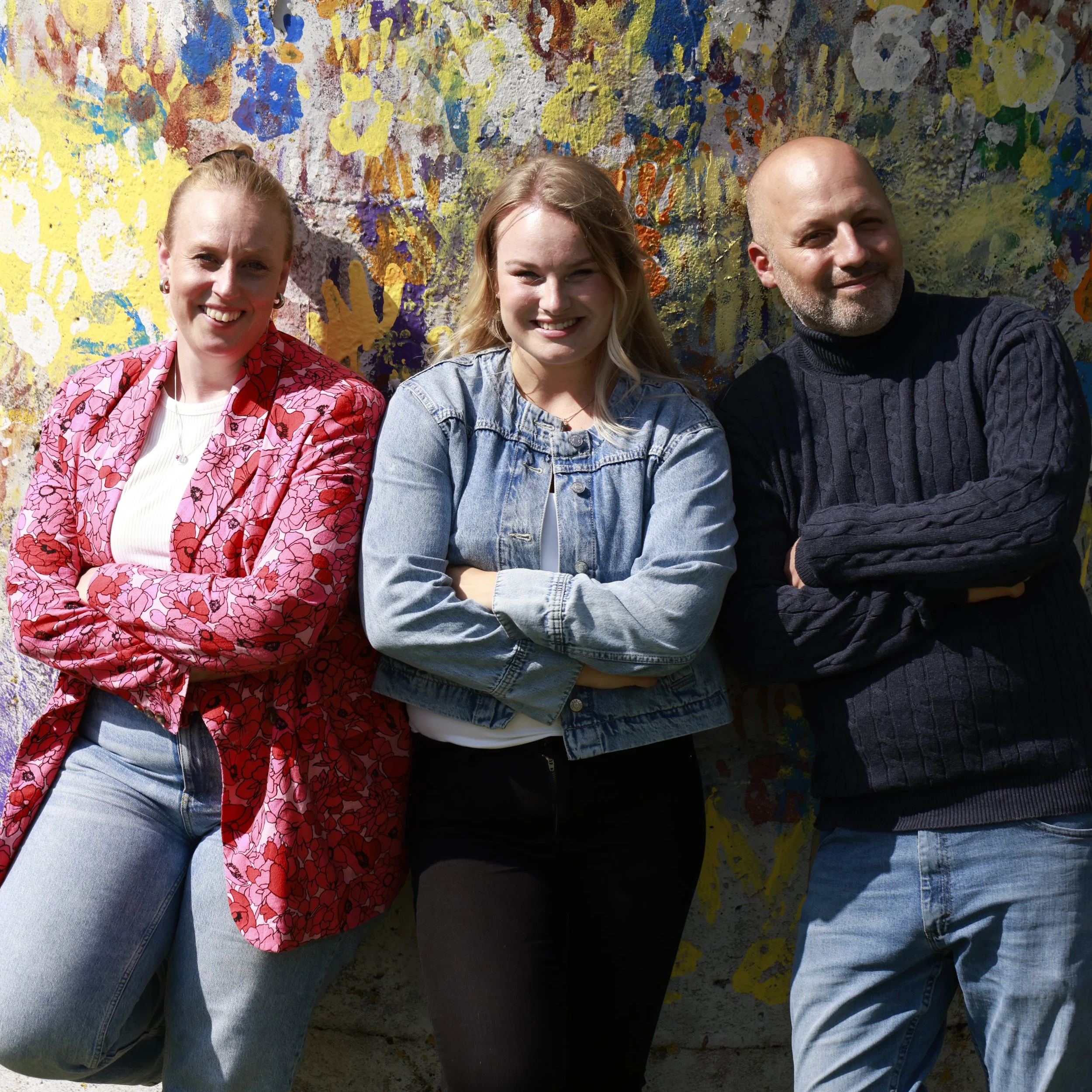 Mikaela, Emelie and Amijaji standing in front of a colorful, abstract mural. Two women and one man are smiling with arms crossed.