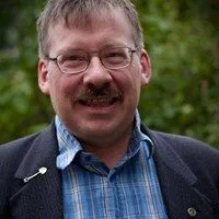 Kenneth with glasses, a mustache, and short hair smiling outdoors with greenery in the background.