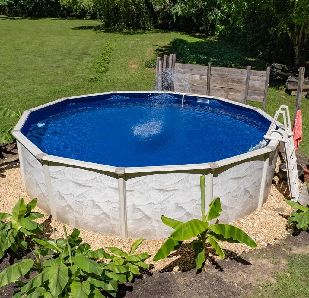 An above-ground octagonal swimming pool filled with blue water in a backyard with green grass and trees, with a ladder on the side and a privacy fence in the background.