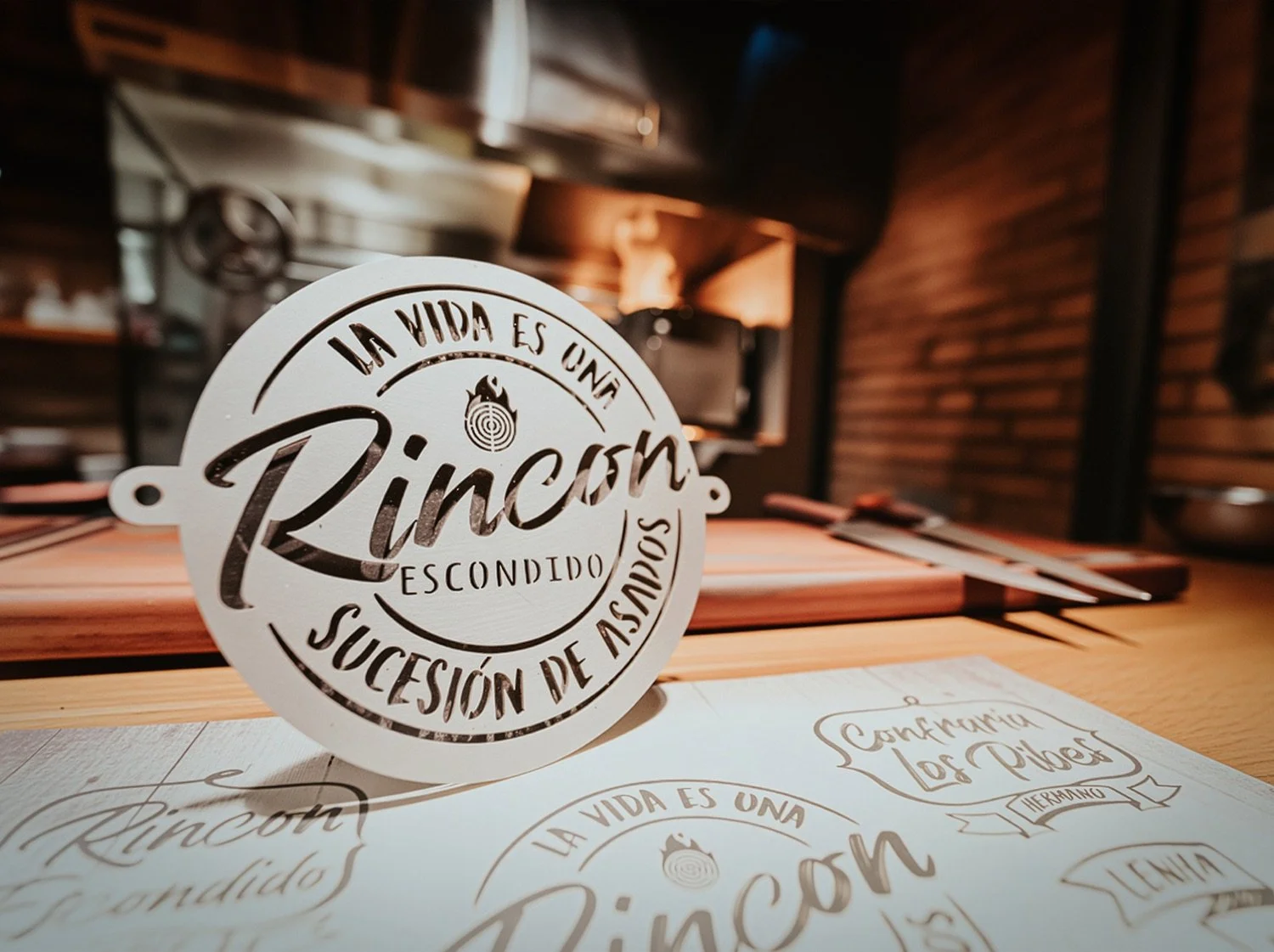 Decorative circular sign with Spanish text in a restaurant setting, featuring the words "La vida es una rincón escondido, sucesión de asombros." The background shows a wooden interior and a flat wooden table with paper menus and utensils.