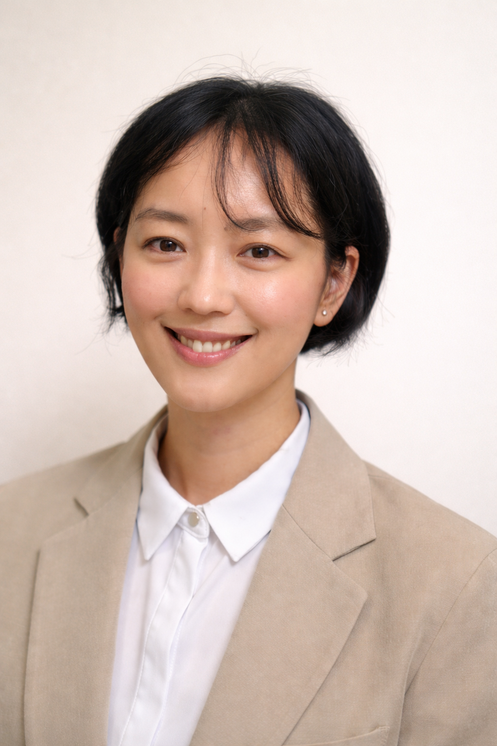 A woman with short black hair, wearing a beige blazer and a white collared shirt, smiling at the camera against a plain background.