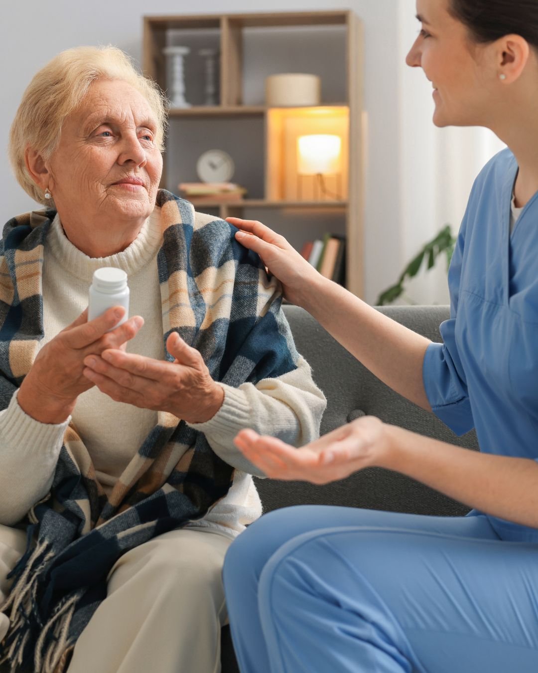 A caregiver in blue scrubs gently touches the shoulder of an elderly woman who is seated on a couch and holding a bottle of medication, with a warm and comforting environment in the background.