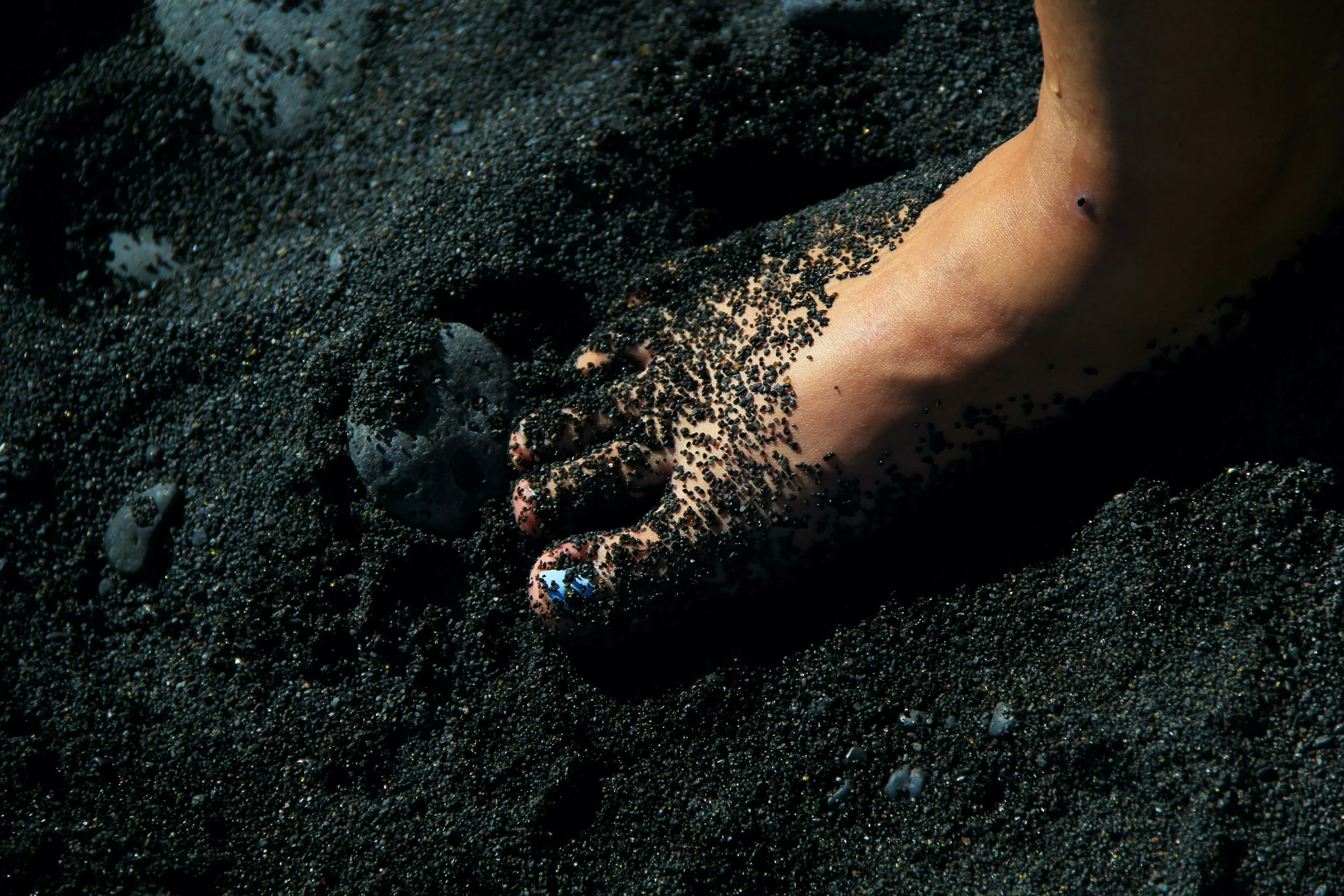 A hand partially buried in dark black sand, with black sand covering fingers and part of the wrist, near a small volcanic rock.