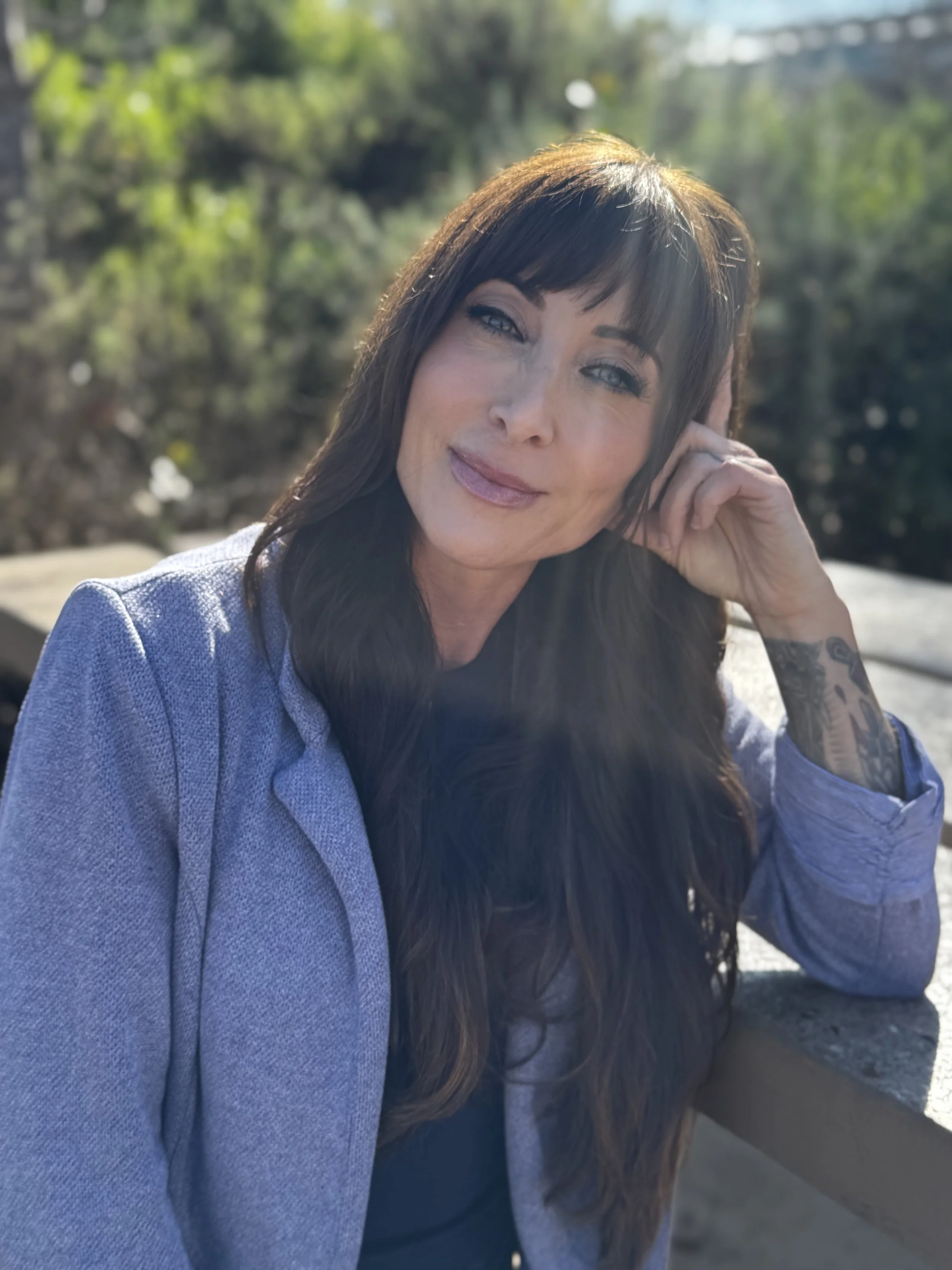 A woman with long dark hair and tattoos, wearing a light blazer, sitting outdoors with greenery in the background, smiling at the camera.