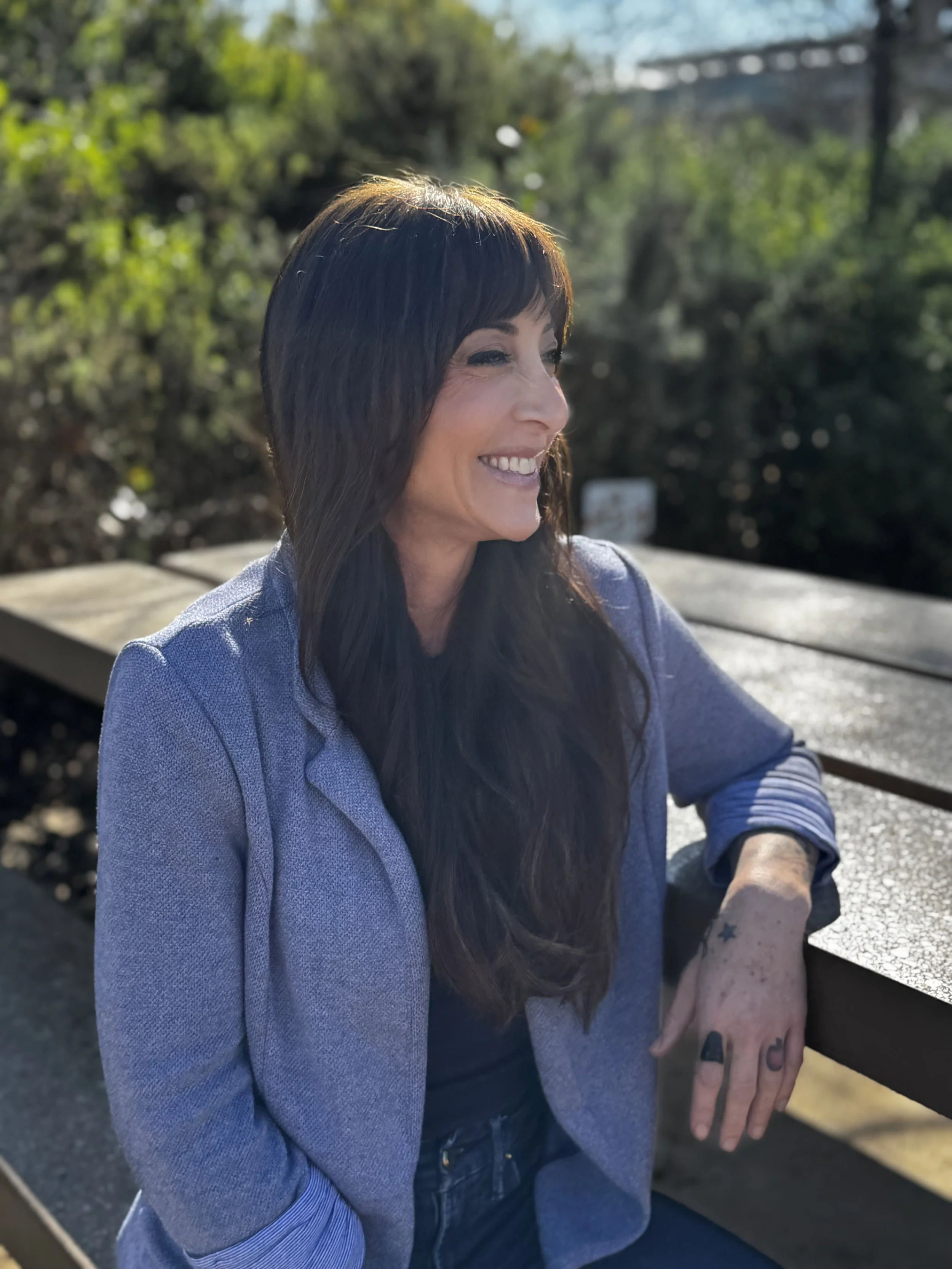 A woman with long dark hair is sitting outdoors on a bench, smiling and looking to the side, with a background of greenery and trees in bright sunlight.