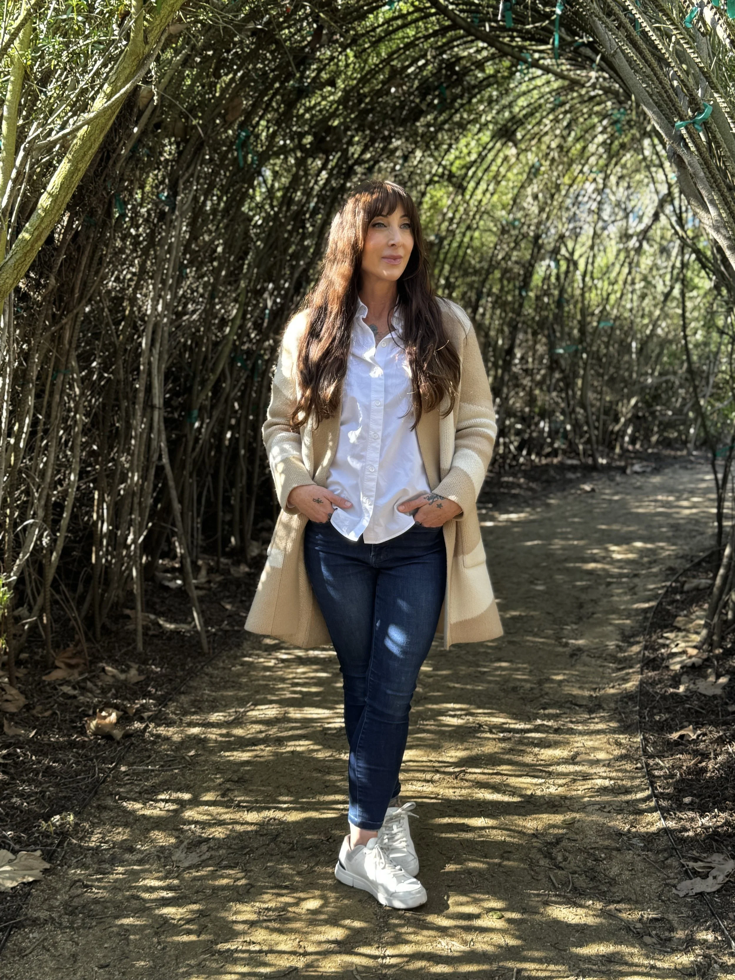 A woman with long brown hair, wearing a white button-up shirt, beige coat, dark jeans, and white sneakers, standing in a tunnel of intertwined branches with greenery overhead, on a dirt path with sunlight filtering through.
