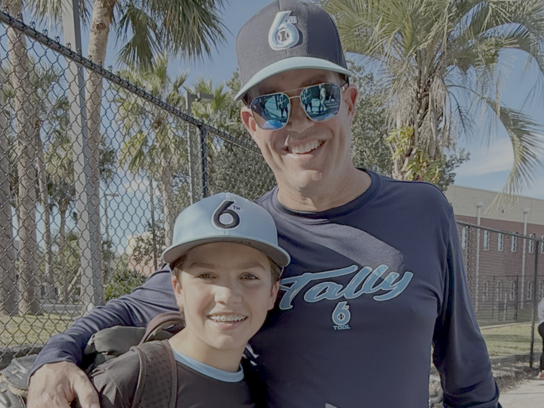 A man and boy smiling outdoors, both wearing baseball caps, with a chain-link fence and palm trees in the background.