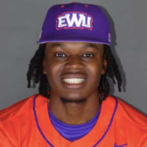 A young man with dark hair in dreadlocks wearing an orange sports jersey, a purple cap with white letters, and smiling showing white teeth against a gray background.