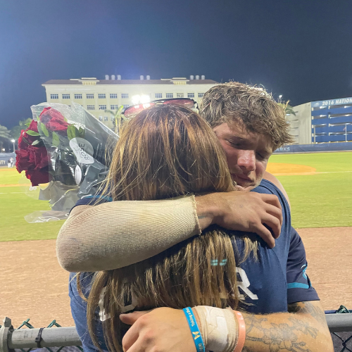 A young man and woman hugging at a baseball stadium during night, with the woman holding a bouquet of red roses.