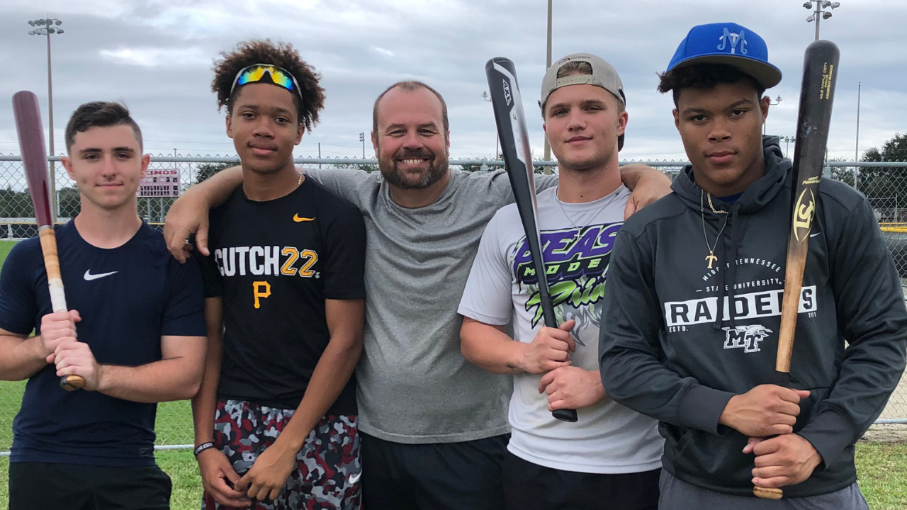 Group of four young male baseball players and a coach standing on the baseball field with their arms around each other, holding baseball bats, and smiling at the camera.