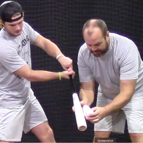 Two men in gray t-shirts and shorts are practicing tennis with a ball launcher on an indoor court. One man is holding the launcher, and the other is holding a tennis ball.