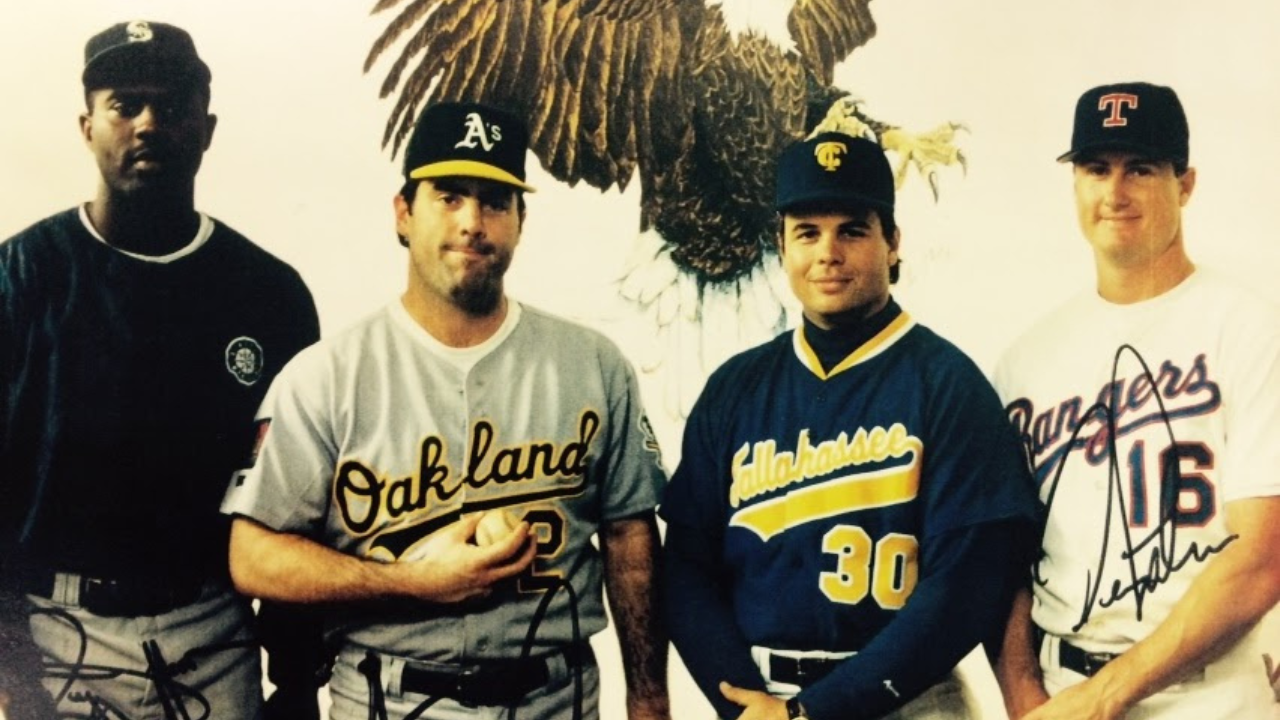 Four young men wearing baseball jerseys and caps standing in front of a wall featuring a large eagle mural.