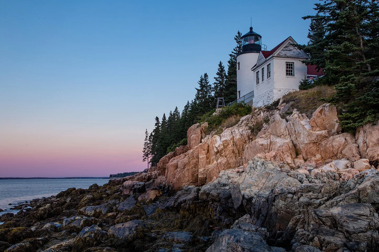 A lighthouse on a rocky shoreline at dusk, with a forested hill and pink and blue sky in the background.
