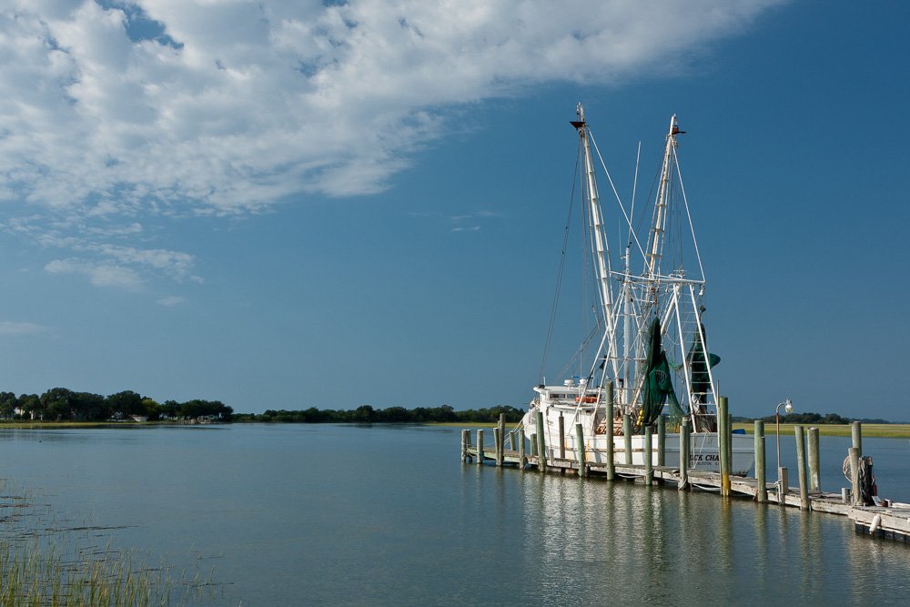 A shrimp boat docked at a pier on calm water with a partly cloudy sky. Photography Classes in Charleston SC. Camera Classes in Charleston SC